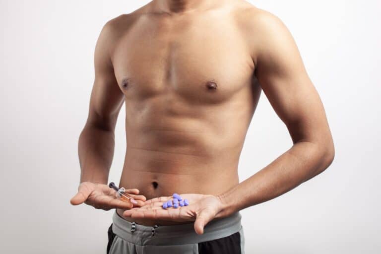 A shirtless muscular man holding hormone treatment syringe and pills on palm